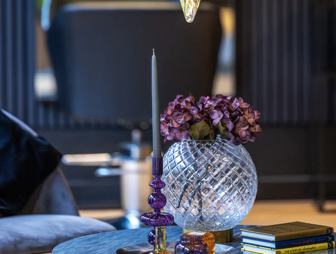 Loungehörna i salongen med glasvas och lila blommor, elegant ljusstake och designböcker på soffbord.